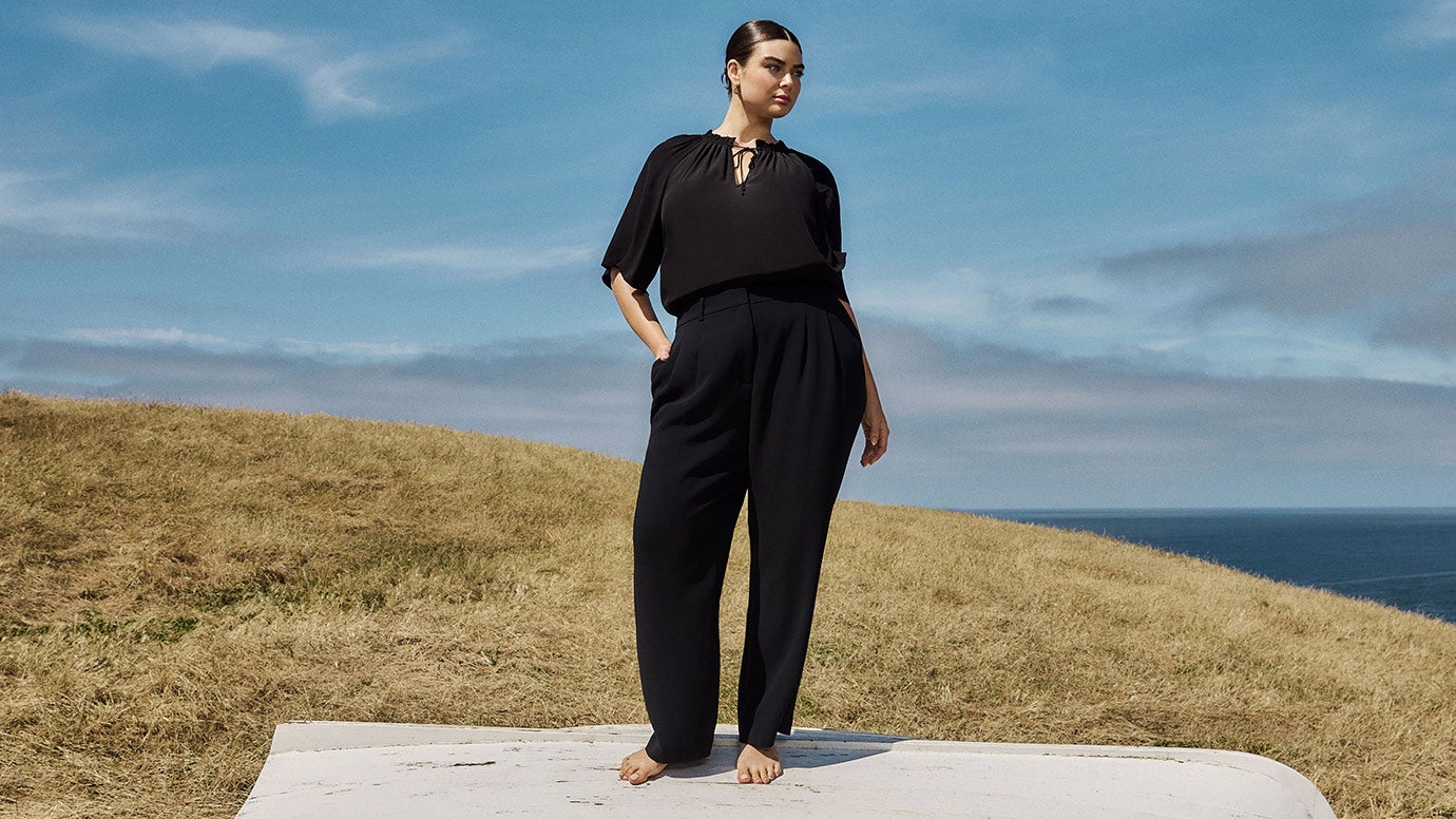 plus size model with dark hair wearing a short sleeve black blouse and black pants standing barefoot on the bottom of an upturned old boat, the setting is a grassy cliff