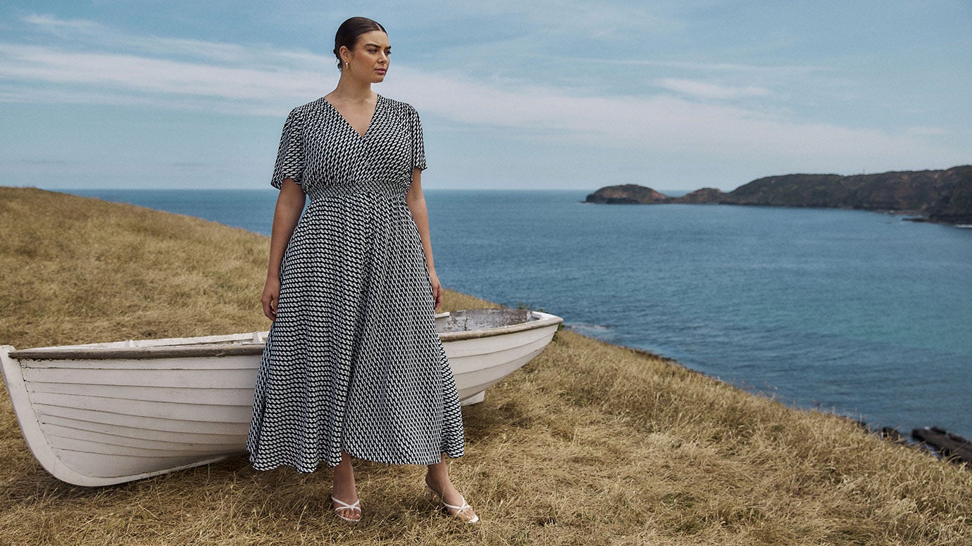 plus size model with slick back dark hair wears a blue and white geo print maxi dress with wrap style bodice, she stands on a cliff looking out to the ocean, behind her is an whitewashed vintage boat