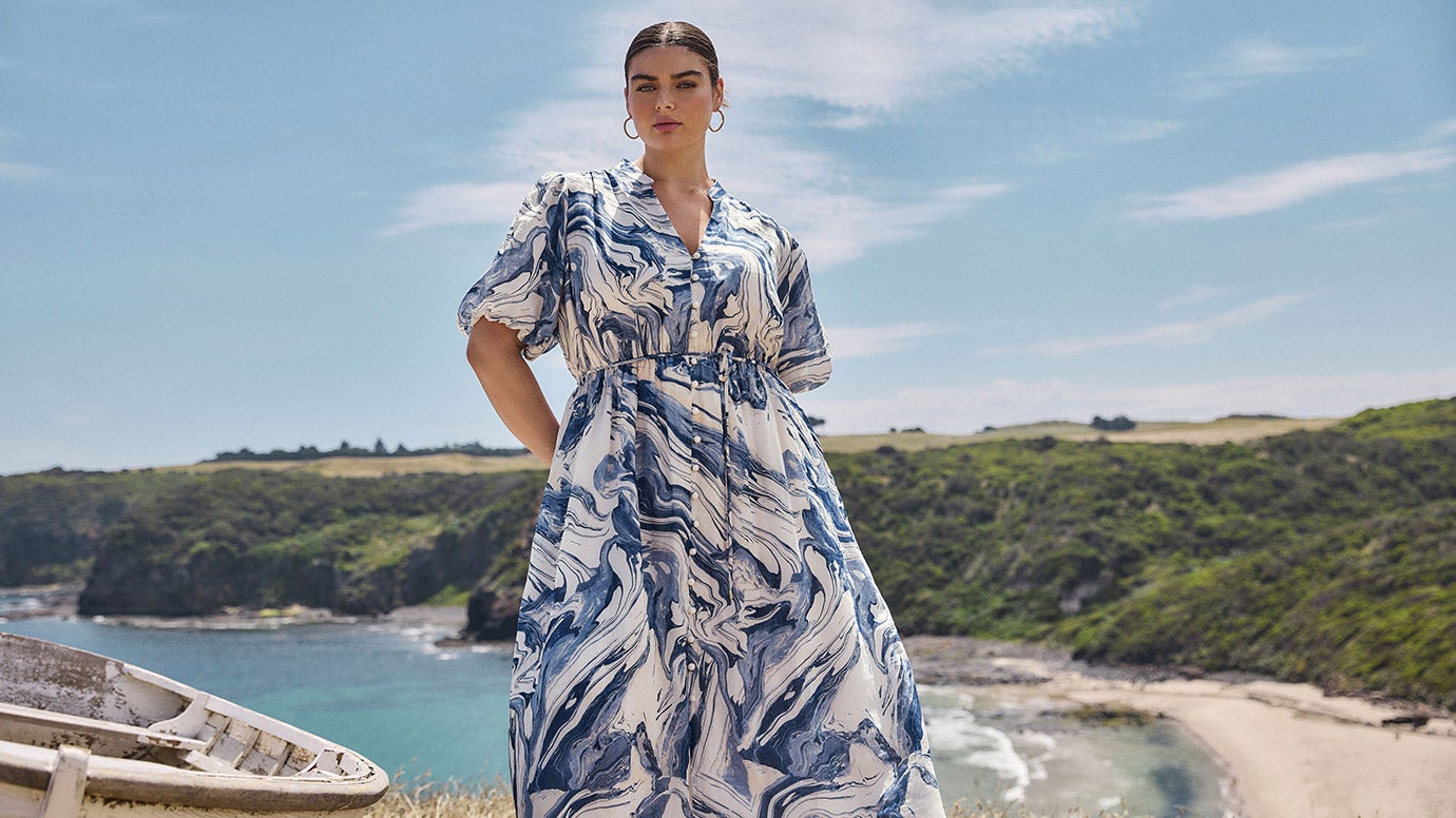 plus size model with dark hair slick back wearing a blue and white marble print dress with puffy sleeves, you can see a beautiful bay or inlet behind with blue water