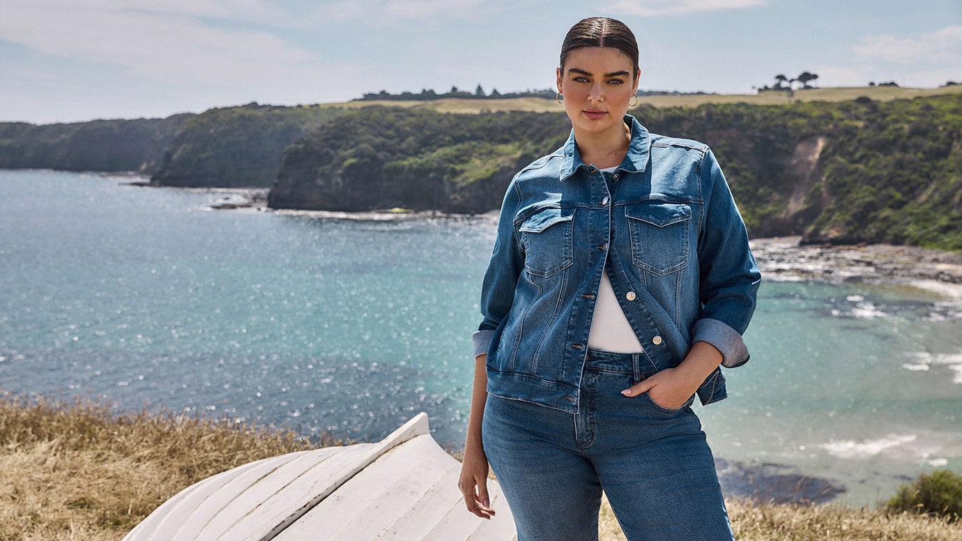 plus size model wears a blue denim jacket and matching jeans, you can see an old boat and the glistening blue ocean behind her