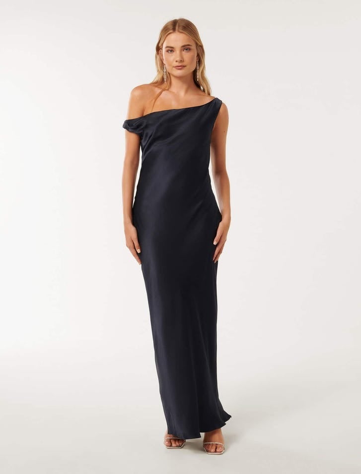 model with long blonde hair standing front on, she is wearing a long deep navy satin dress with a draped on shoulder design, she is wearing strappy heels