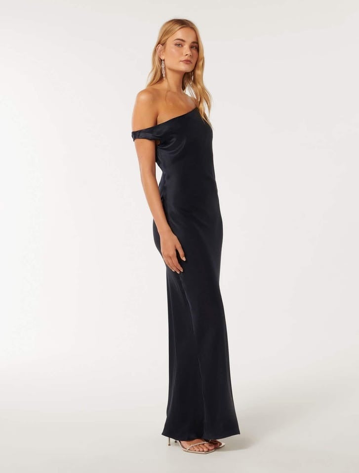 model with long blonde hair standing in profile but with her head turned toward the camera, she is wearing a long deep navy satin dress with a draped on shoulder design, she is wearing strappy heels